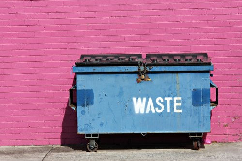 A large blue industrial waste bin with the word 'WASTE' printed in white on its front, positioned on a concrete pavement against a vibrant pink painted brick wall. The bin appears to be made of metal with a slightly worn surface, and features a hinged lid secured by a metal latch at the top. The wheels underneath suggest ease of movement, and the surrounding environment indicates an outdoor area possibly associated with commercial waste collection in Harrow. The lighting suggests natural daylight, casting a shadow of the bin onto the pavement, emphasizing its rectangular shape and the texture of the metal surface. This scene aligns with professional rubbish removal services provided by Commercial Waste Harrow, serving local businesses needing waste management solutions.