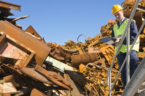 A male waste management inspector wearing a yellow safety helmet and high-visibility vest stands on a metal staircase at a scrap yard, examining a clipboard while faced with a large pile of rusted metal debris. The debris includes various sheets, beams, and industrial-grade metal parts, exhibiting a rough, weathered texture with shades of orange, brown, and dark grey, indicating corrosion and age. The background features a clear blue sky and a small portion of trees with autumn foliage, suggesting a daytime outdoor setting near Harrow. The scene emphasizes the process of rubbish management and inspection, with the rusted metal primarily occupying the foreground and middle ground, while the inspector is positioned on the right side of the image, observing the site attentively. The environment appears to be an industrial or recycling site, with the large pile of waste materials indicating active rubbish removal activities consistent with services provided by Commercial Waste Harrow.