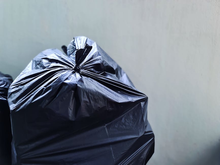 Close-up of a large black plastic rubbish bag tightly knotted at the top, placed against a plain, light-colored wall. The bag is made of shiny, flexible polyethylene material with visible creases and